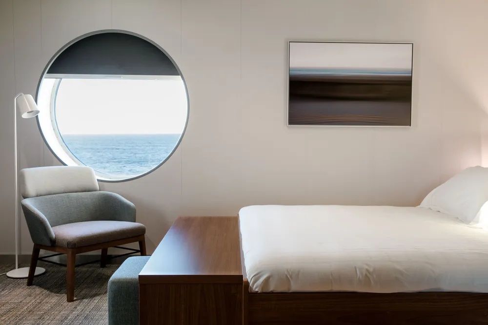 Camarote moderno a bordo de un ferry, con cama doble, zona de estar y una ventana circular con vistas al mar.