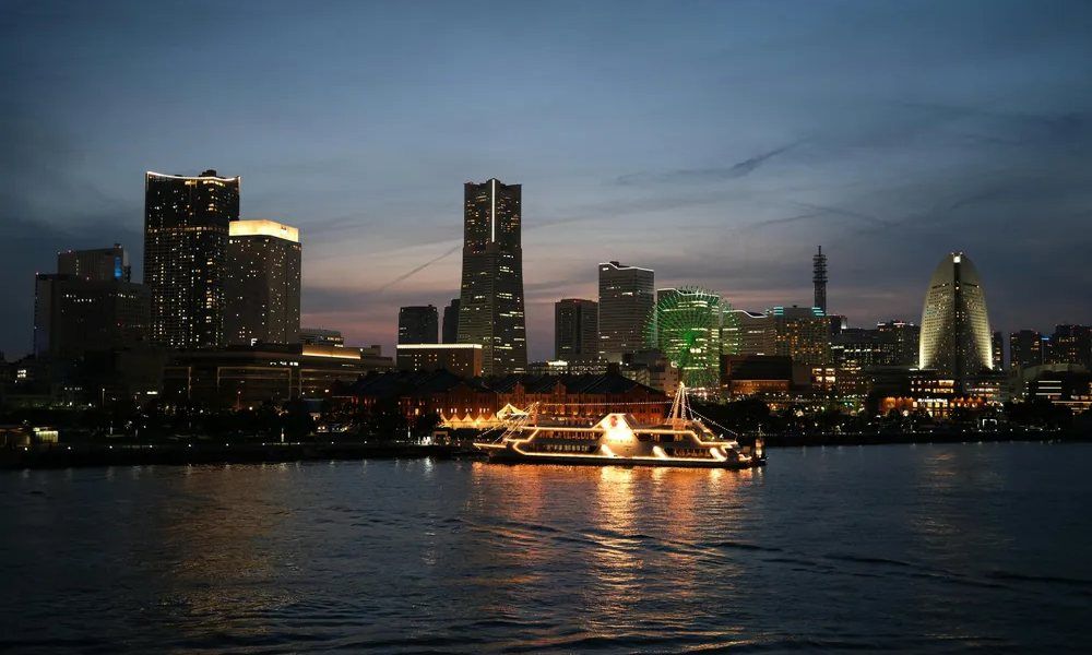 Panorámica nocturna de una ciudad junto al agua, con edificios iluminados y un barco con luces navegando en primer plano.