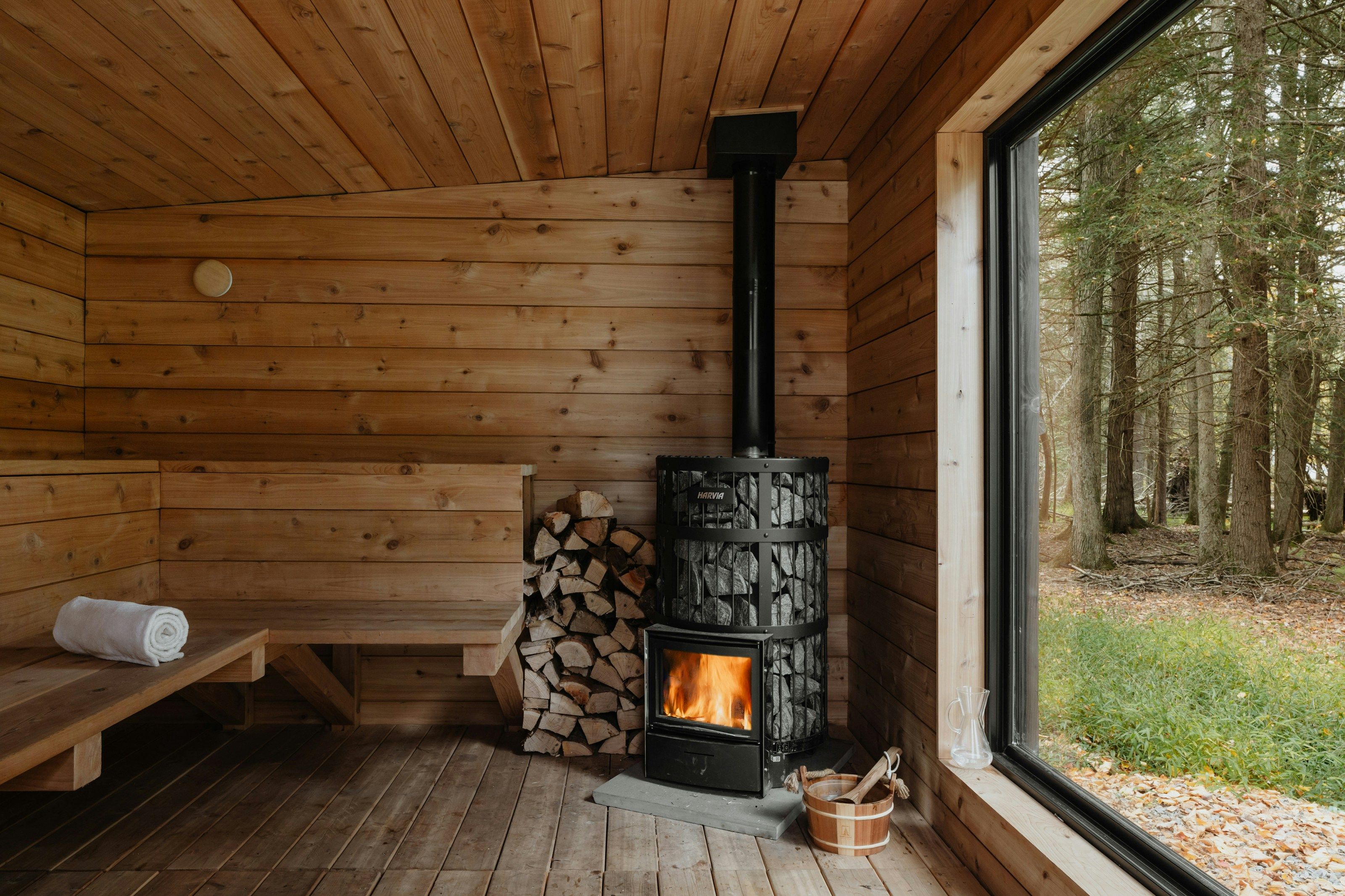 Eine leere finnische Sauna von innen. Sie ist aus Holz, links ist die Sitzbank und ein gerolltes Handtuch. Mittig sieht man den brennenden Kamin, einen Stapel Holz und einen Aufguss-Eimer. Rechts ist ein großes Fenster mit Ausblick auf einen Wald.