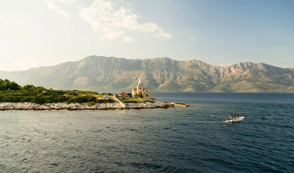 Pequeño faro y edificio en una isla del Adriático con montañas al fondo y bote navegando.
