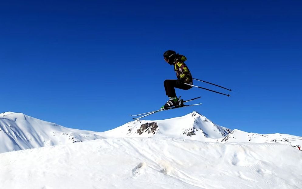 Esquiador realizando un salto en una pista nevada bajo un cielo azul despejado.