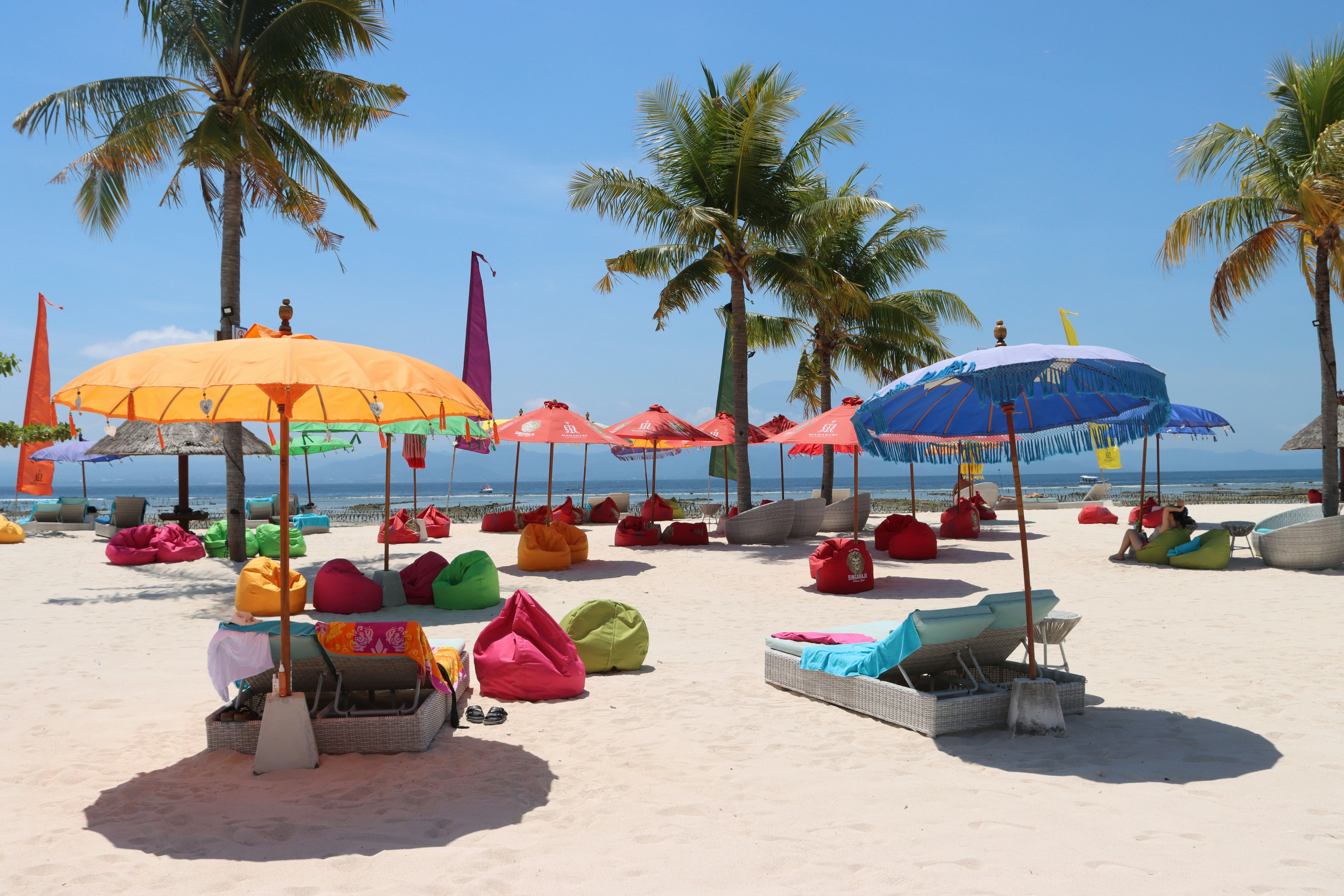 Am weißen Sandstrand von Nusa Lembongan in Indonesien stehen farbenfrohe Sonnenschirme mit Liegen. Im Hintergrund sieht man Palmen.