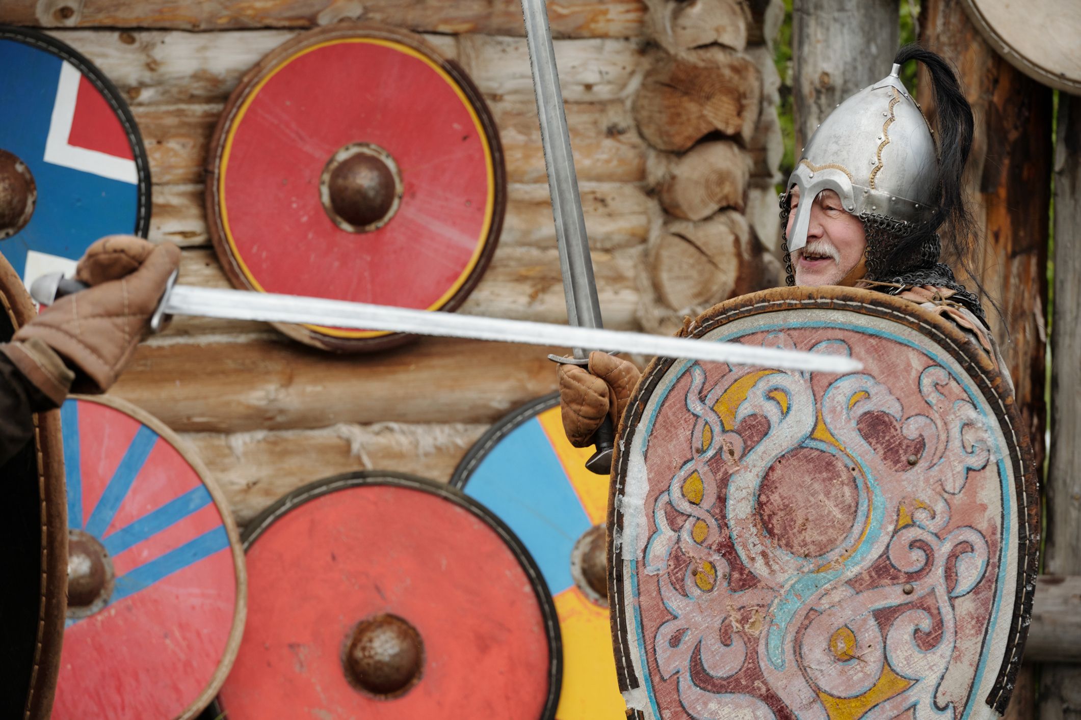 Ein älterer Herr trägt einen Wikinger-Helm, in einer Hand hat er ein Schwert, in der anderen ein Schild. Eine weitere Person hält ein Schwert auf das Schild. Der ältere Herr lacht.