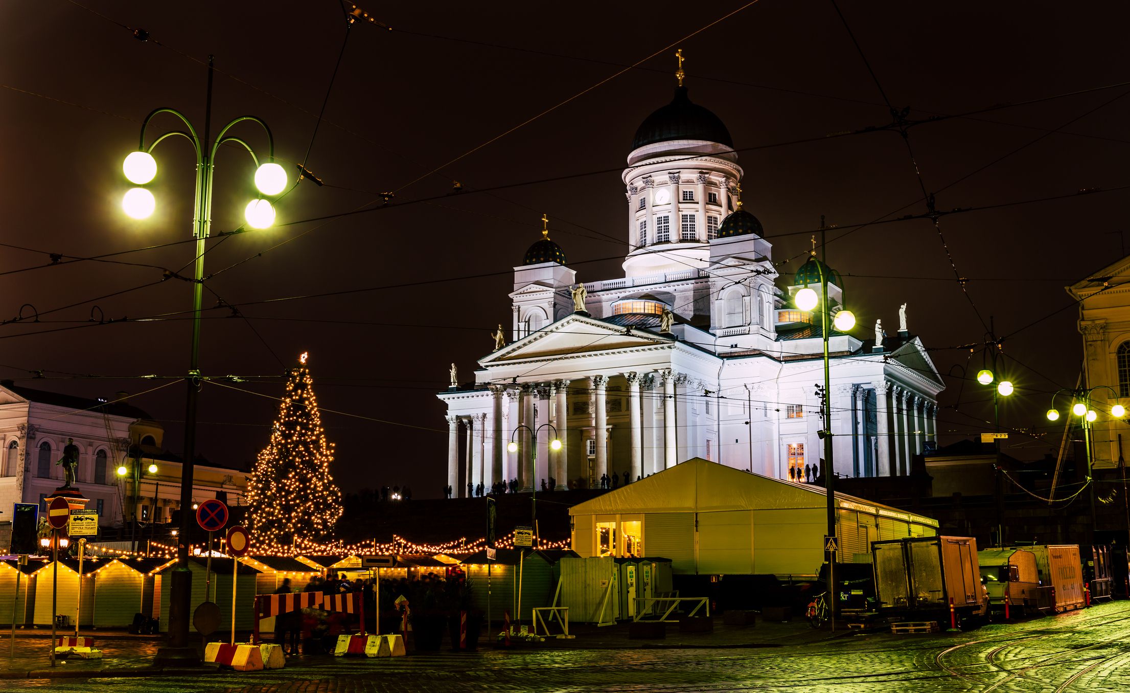 Der Dom von Helsinki ist bei Nacht angestrahlt. Im Vordergrund ist der festliche Weihnachtsmarkt mit dem bunt beleuchteten Weihnachtsbaum.