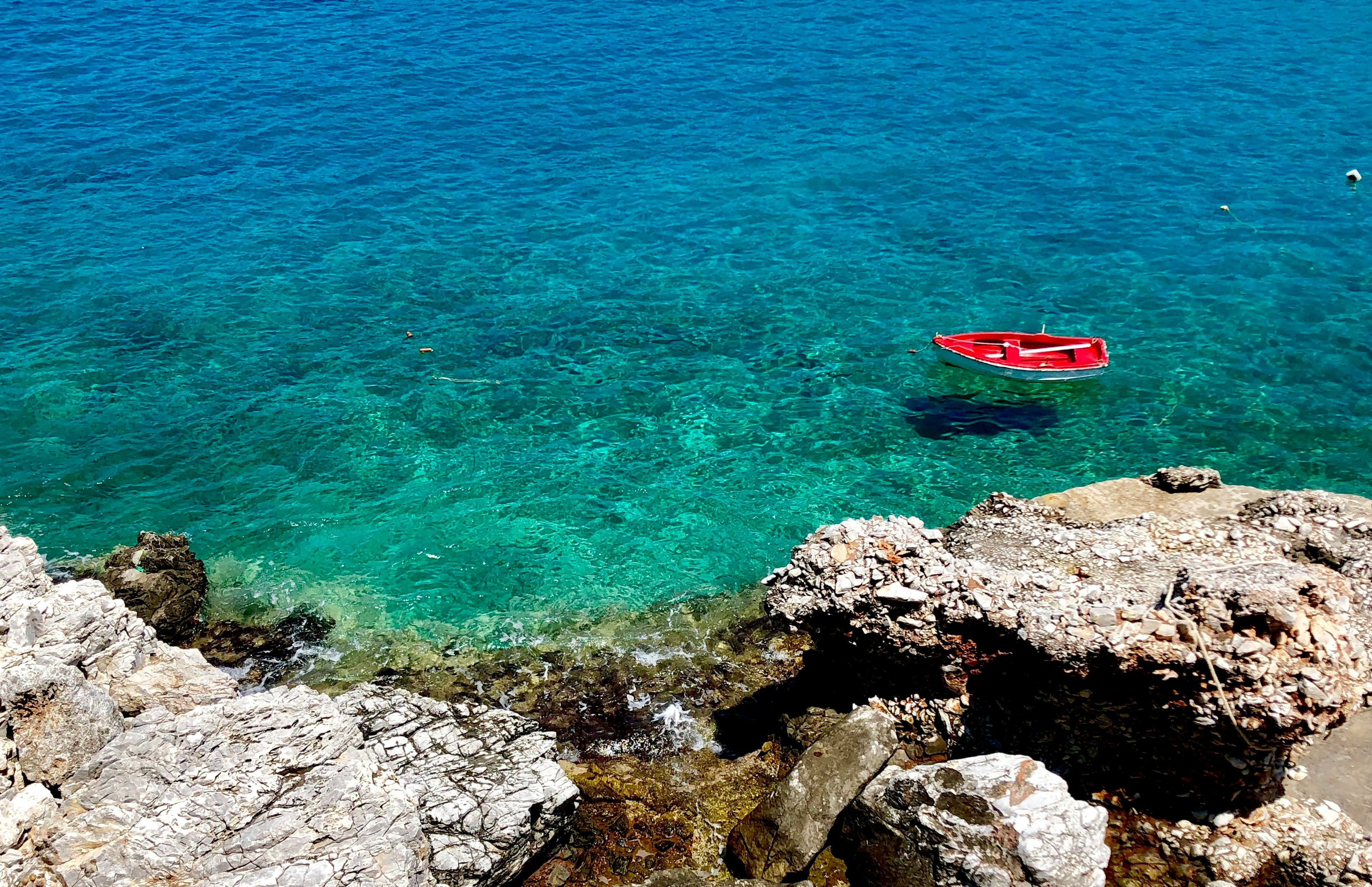 Steinklippe im Vordergrund, klares blaues Meer auf dem ein kleines rotes Boot schwimmt.