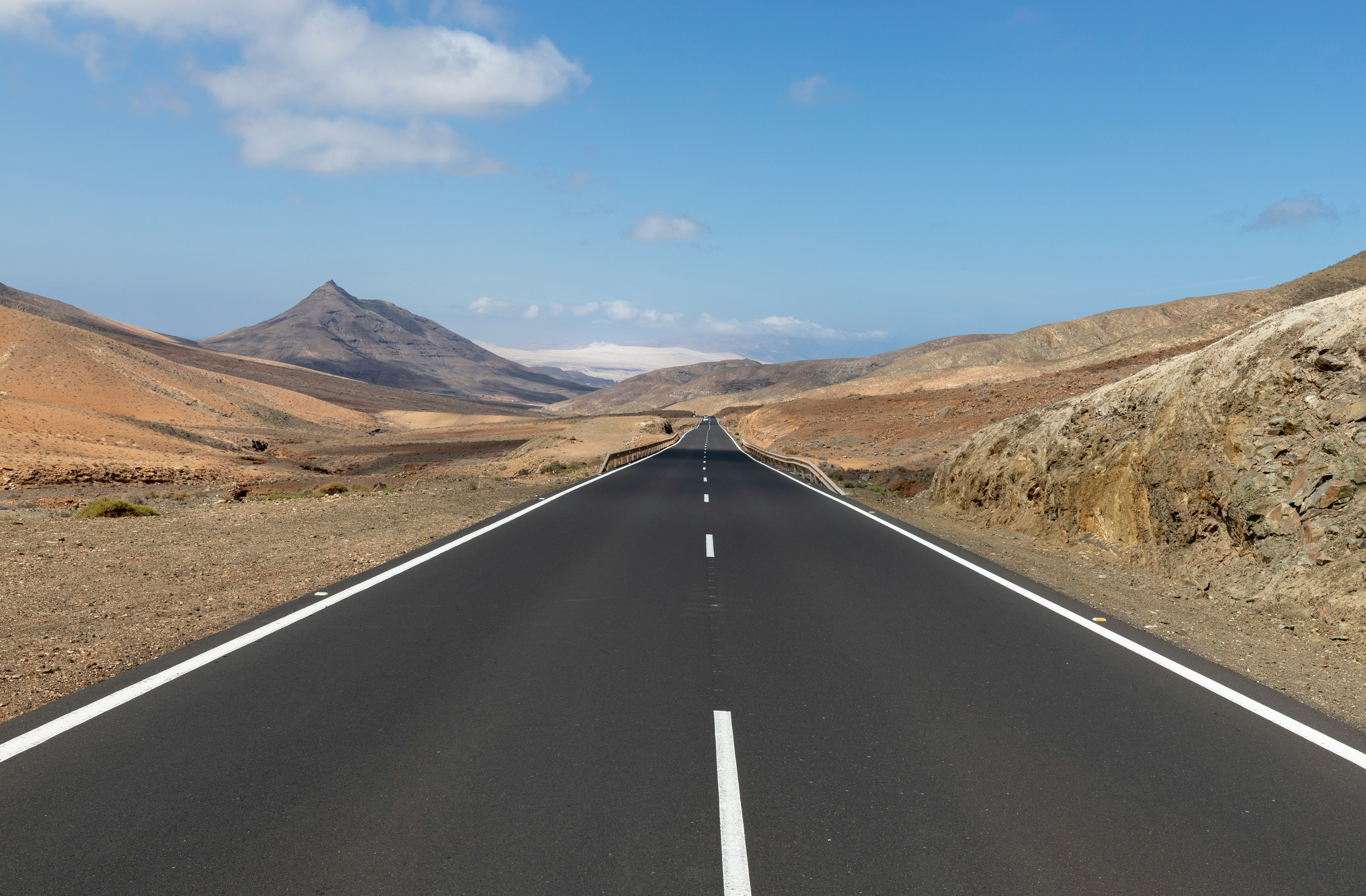 Eine leere asphaltierte Straße ist im Fokus des Bildes und führt in den Hintergrund. Links und rechts ist karges Land, im Hintergrund sieht man einen Berg.