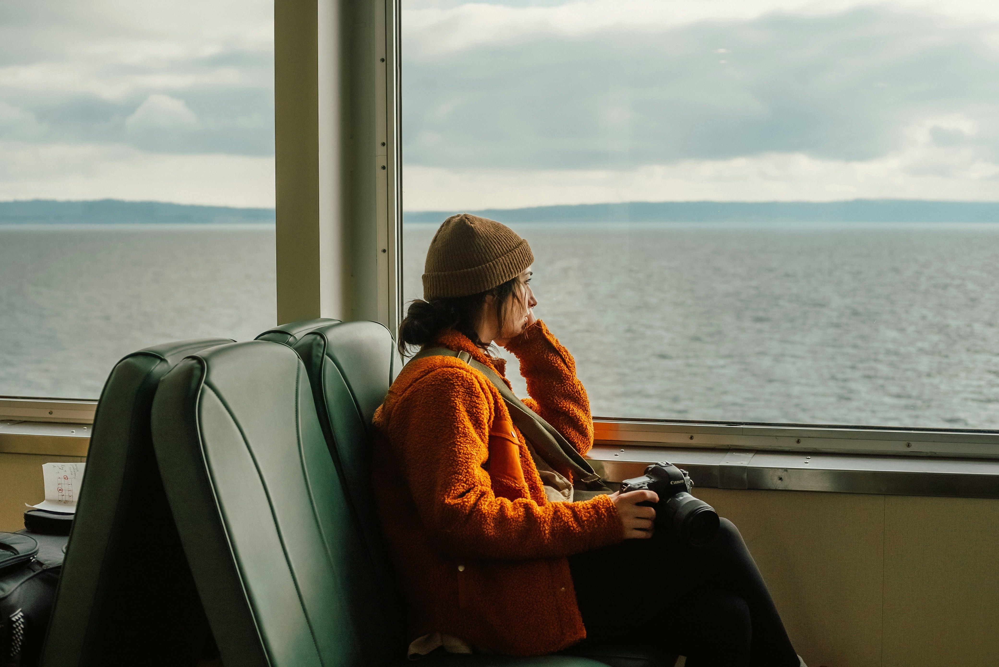 Een vrouw zit op een groene bank op een veerboot en kijkt door het grote raam naar de zee. Ze draagt ​​een bruine hoed, een oranje jas en een donkere broek, en heeft een camera aan een riem om haar nek hangen.
