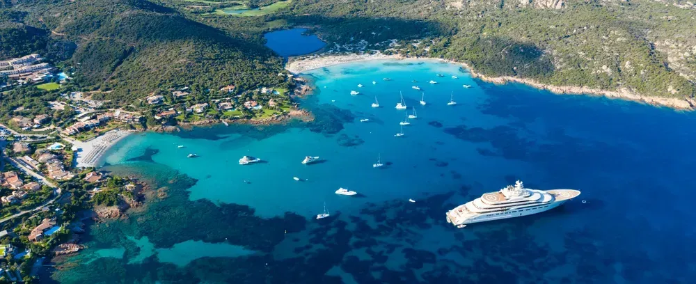 Eine Luftaufnahme der exklusiven Costa Smeralda auf Sardinien. Auf dem tiefblauen Wasser treiben einige kleinere Boote sowie eine Superyacht.