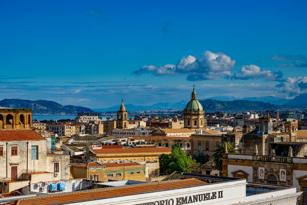 Die Dächer von Palermo strahlen kräftig in der Sonne und unter einem blauen Himmel. Im Hintergrund kann man das Meer erahnen.