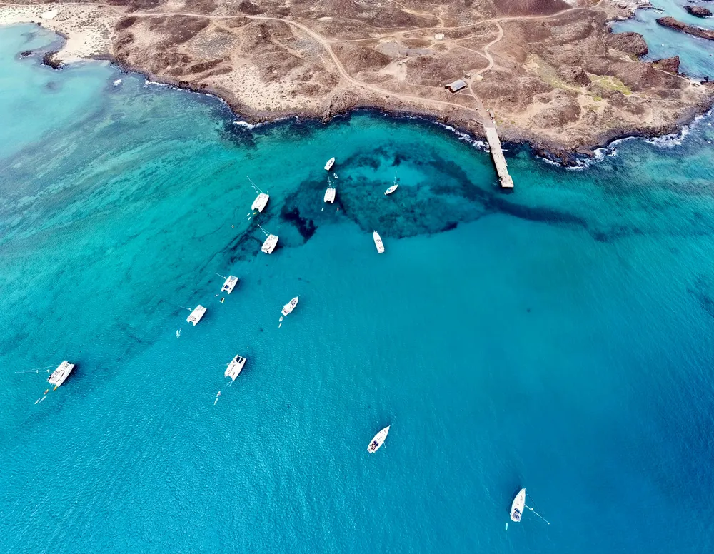 Eine Luftaufnahme der Küste von Isla de Lobos. Im blauen Meer treiben ein paar Ausflugsboote, im oberen Bereich des Fotos sieht man den Anlegesteg und die Küste der Insel.