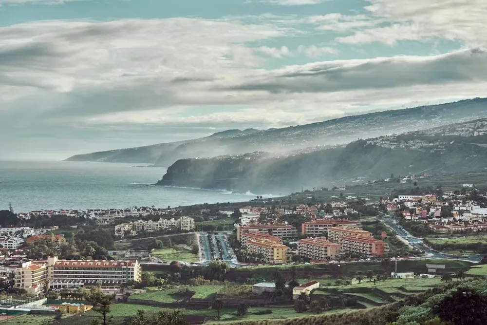 Vista elevada de la costa norte de Tenerife: urbanizaciones y hoteles de baja altura rodeados de campos verdes, con acantilados que se pierden en la bruma marina y montañas al fondo.