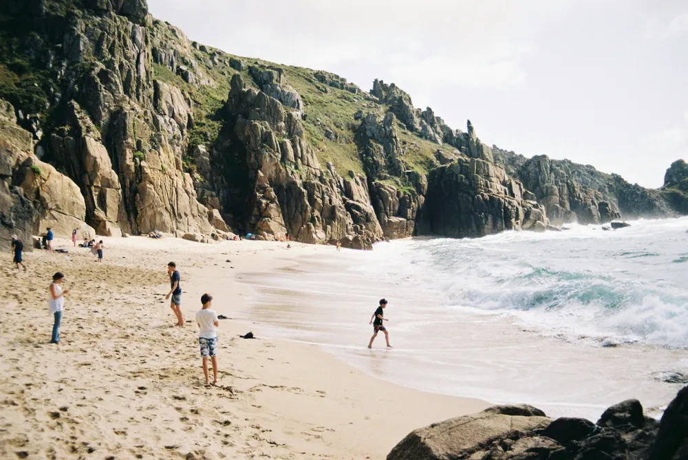 Een paar kinderen spelen op het strand. Links op de foto is het zand, rechts de zee. Op de achtergrond zijn rotsen te zien.