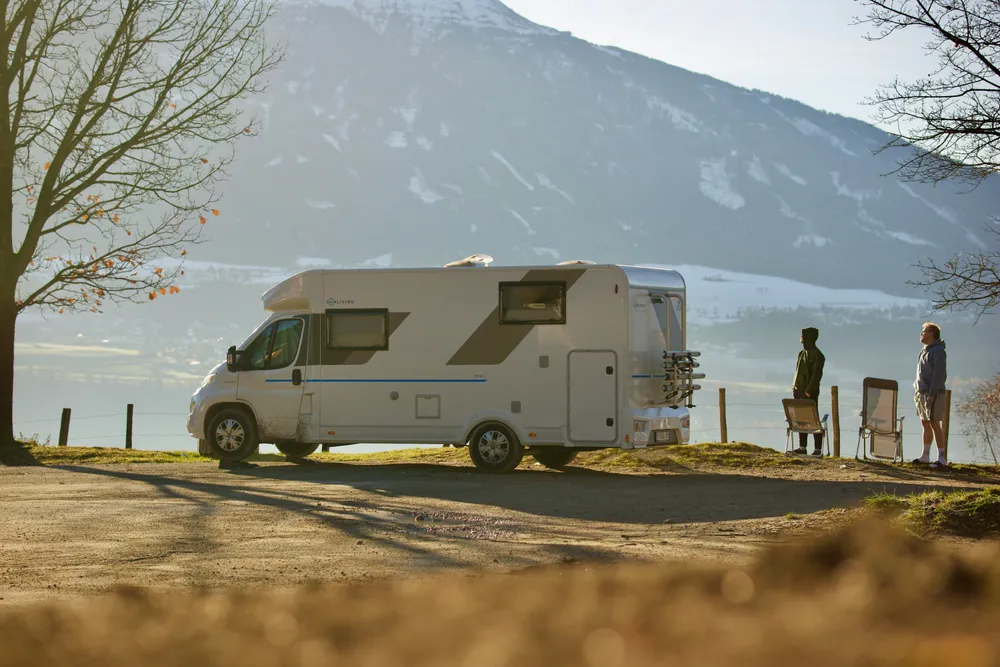 Ein Wohnmobil steht vor einem leicht mit Schnee bedeckten Berg. Die Sonne scheint leicht von links ins Bild, rechts vom Wohnmobil stehen zwei Menschen.