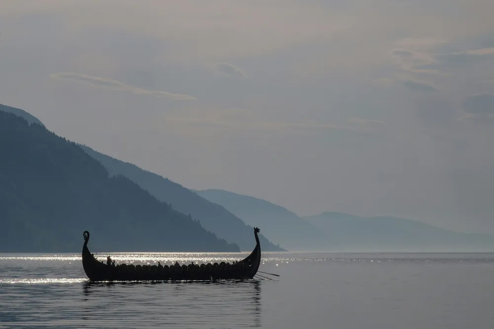 Ein Nachbau eines Langschiff der Wikinger gleitet über das ruhige Wasser. Im Hintergrund kann man eine Fjord-Landschaft erahnen.