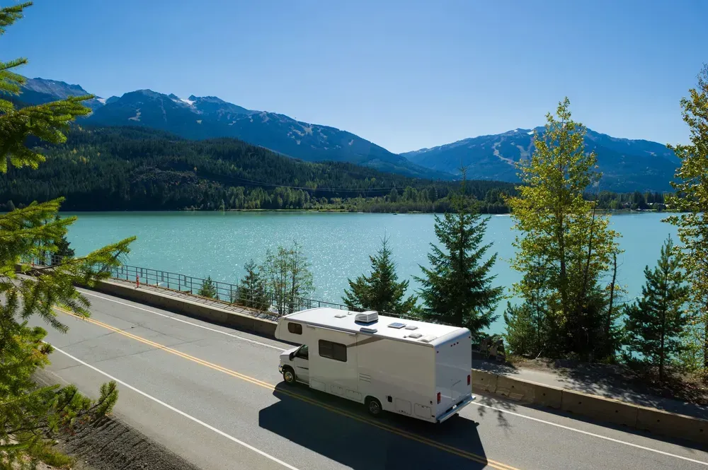 Een witte camper rijdt langs een schilderachtig meer. De zon schijnt, het turquoise water glinstert. Op de achtergrond zijn dichte, groene bossen te zien.