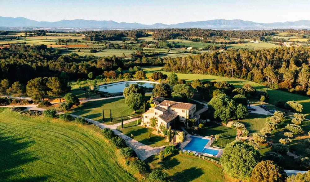 Vista aerea di una fattoria rurale nella Garrotxa circondata da campi verdi, alberi e un piccolo lago, con le montagne di Girona sullo sfondo.