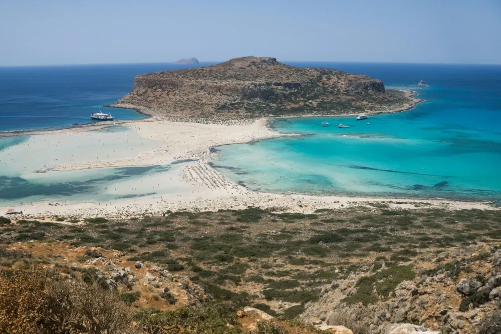 Das Foto wurde von einem Hügel aus aufgenommen und zeigt einen Blick auf einen Strand. Über das offene Meer hinaus erkennt man eine Insel.