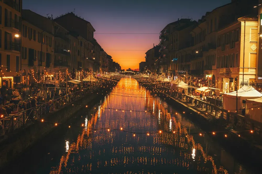 Ein Kanal in Mailand bei Nacht. Im Wasser werden Lichter reflektiert, die aus den links und rechts entlang laufenden Gaststätten und Gebäuden kommen. Über dem Wasser hängen Lichterketten.