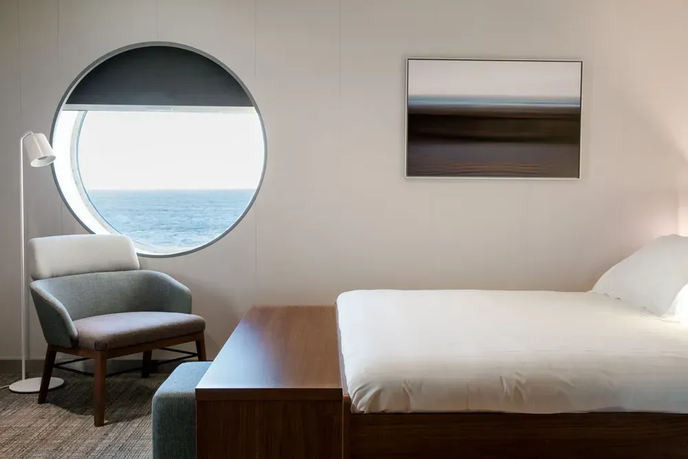 Een foto van een hut op een veerboot. Een comfortabel bed met wit beddengoed is zichtbaar, en daarboven hangt een landschapsschilderij. Links van de foto staan ​​een fauteuil, een staande lamp en een rond raam met uitzicht op zee.