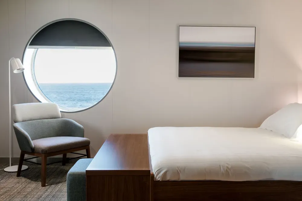 Ein Foto von einer Kabine auf einer Fähre. Man sieht ein komfortables Bett mit weißer Bettwäsche, darüber hängt ein Landschaftsbild. Links im Bild ist ein Sessel, eine Stehlampe und ein rundes Fenster mit Meerblick.