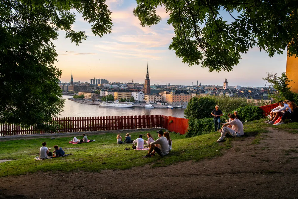Einige Menschen sitzen bei Sonnenuntergang im Sommer auf einem Hügel und schauen aufs Wasser. Im Hintergrund sieht man die Skyline von Stockholm, Schweden.
