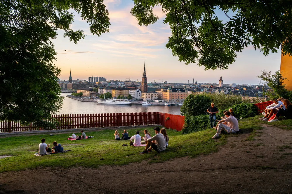 Enkele mensen zitten bij zonsondergang in de zomer op een heuvel en kijken uit over het water. Op de achtergrond zie je de skyline van Stockholm, Zweden.