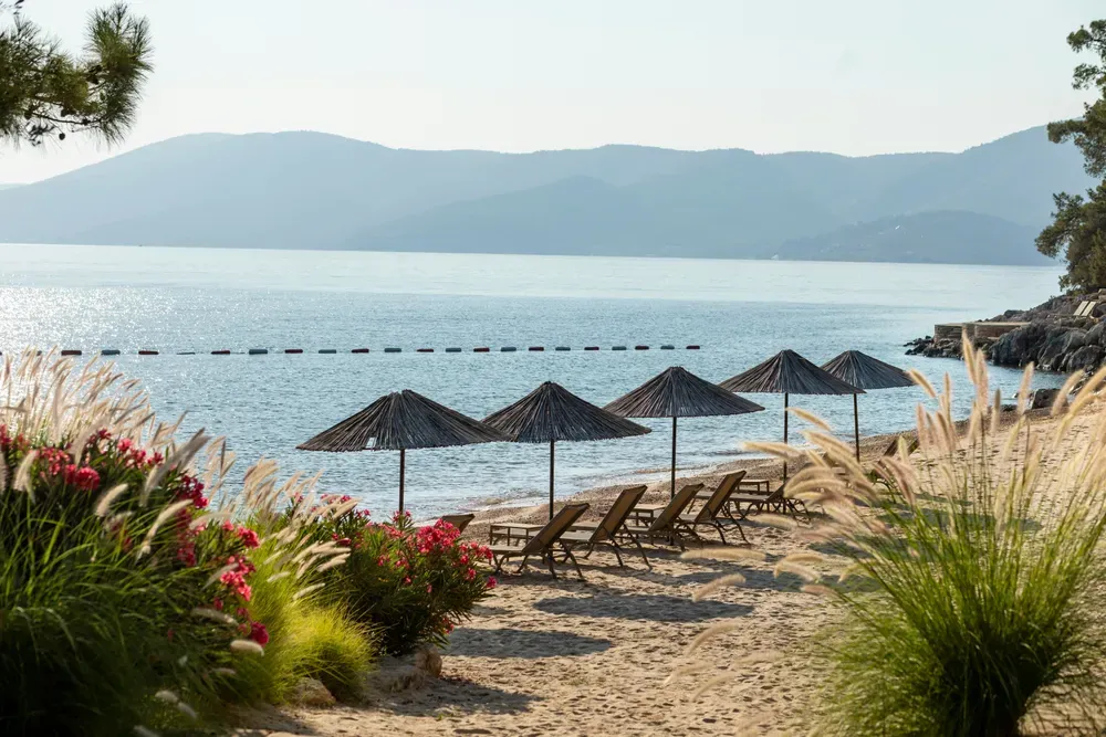 Ein Blick auf einen Strandabschnitt. Im Vordergrund ist Sandstrand mit fünf Stroh-Sonnenschirmen und fünf Sonnenliegen. Im Hintergrund glitzert das Meer in der Sonne, und Berge ragen empor.