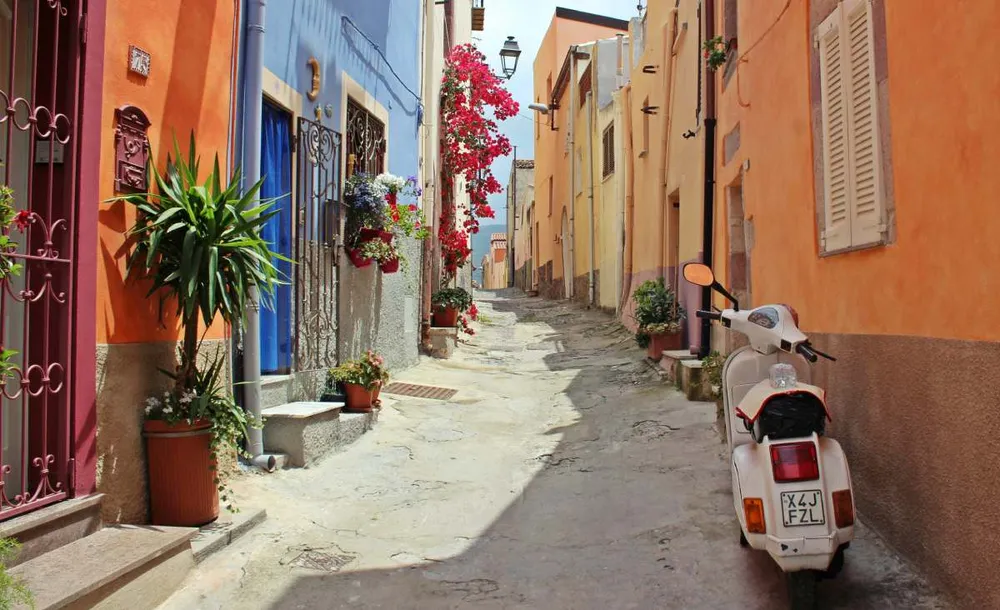 Calle estrecha y colorida en Cerdeña con fachadas tradicionales y una Vespa aparcada, típica de los pueblos italianos.