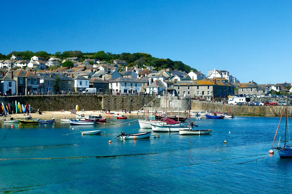 Uitzicht op een baai in Cornwall. Het water is blauw, er liggen een paar boten in het water. Daarachter ligt wat zand, een rotswand en het begin van een klein stadje.