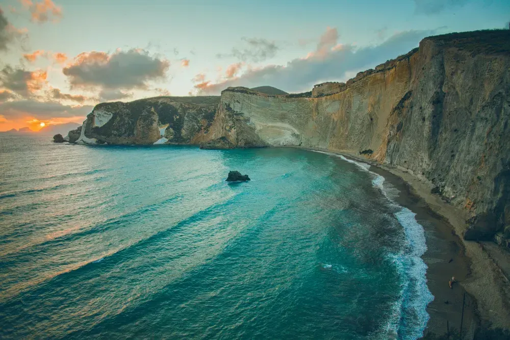 Mächtige Felsklippen winden sich im rechten Bereich des Bildes, links sieht man das Meer mit Schaumkronen am Ufer. Es ist Sonnenuntergang.