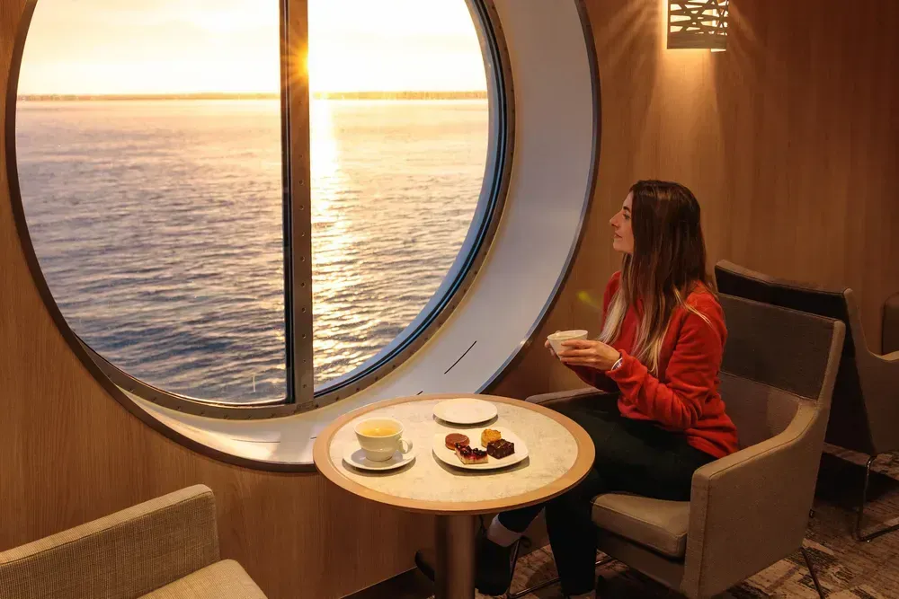 Een vrouw zit bij zonsopgang op een comfortabele stoel op een ferry. Op een ronde tafel voor haar staat een klein ontbijt. Ze houdt een koffiekop vast en kijkt door een groot rond raam uit over zee.