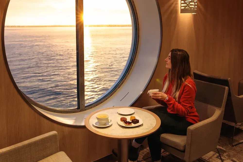 Eine Frau sitzt bei Sonnenaufgang auf einem bequemen Stuhl auf einer Fähre. Auf einem runden Tisch vor ihr steht ein kleines Frühstück. Sie hält eine Kaffeetasse und schaut durch ein großes rundes Fenster aufs Meer hinaus.