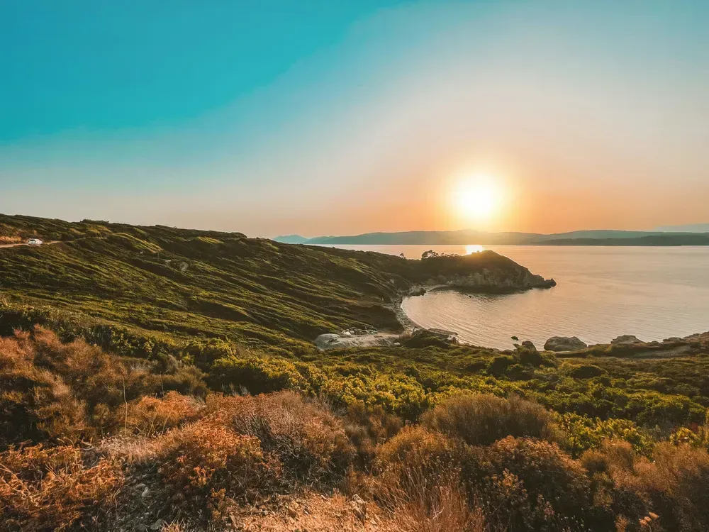 Blick von einem Hügel auf eine halbmondförmige Bucht. Die Landschaft des Hügels ist grün, am Horizont geht die Sonne gerade unter.