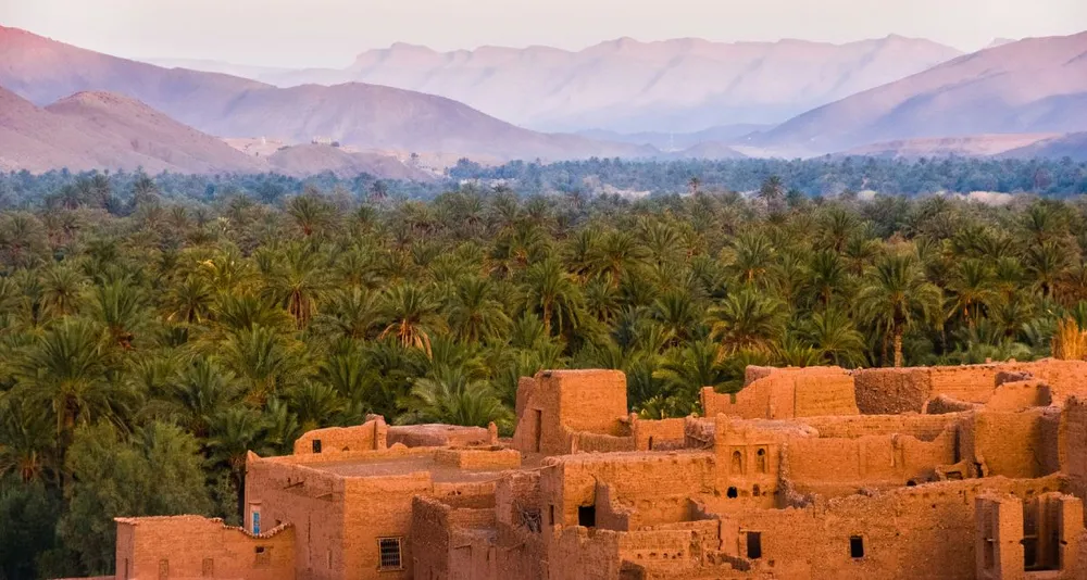 Antiguas casas de adobe en Marruecos junto a un gran palmeral, con montañas al fondo al atardecer.