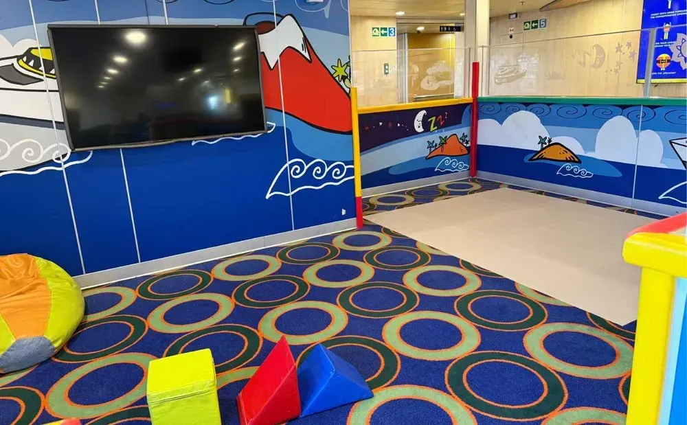 Een kleurrijke speelplek op een veerboot. Tinten blauw, rood en geel creëren een levendige sfeer. Speelgoed ligt verspreid over een speelmat, de muren zijn felgekleurd en aan een van de muren hangt een uitgeschakelde televisie.