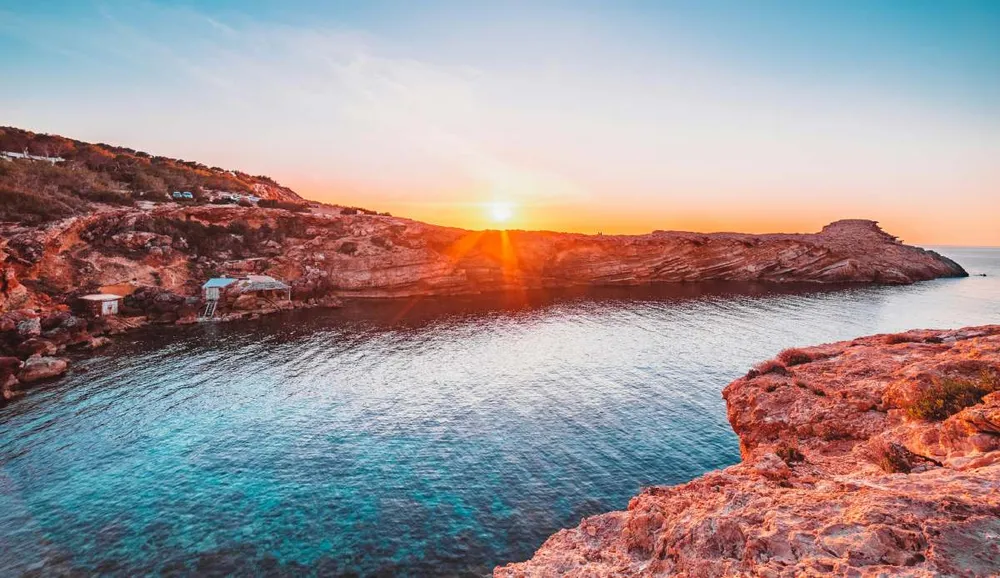 Puesta de sol sobre una cala rocosa en Ibiza, con el mar Mediterráneo iluminado en tonos cálidos.
