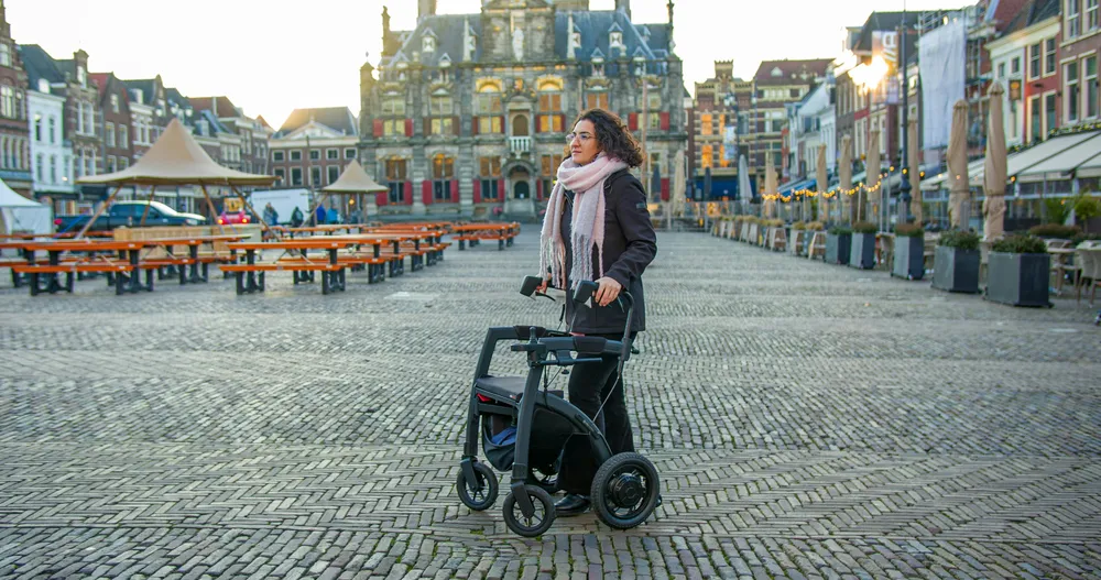 Eine Frau läuft mit einem Rollator über das Kopfsteinpflaster eines Platzes. Sie hat braune Locken und trägt einen pinken Schal.
