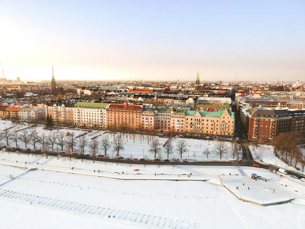 Verschneites Helsinki aus der Vogelperspektive. Im Vordergrund eine verschneite Fläche, im Hintergrund die Häuser der Stadt in der kalten Wintersonne.