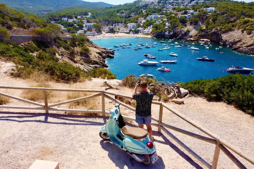 Persona con scooter en un mirador con vistas a una bahía de Ibiza llena de barcos y casas blancas