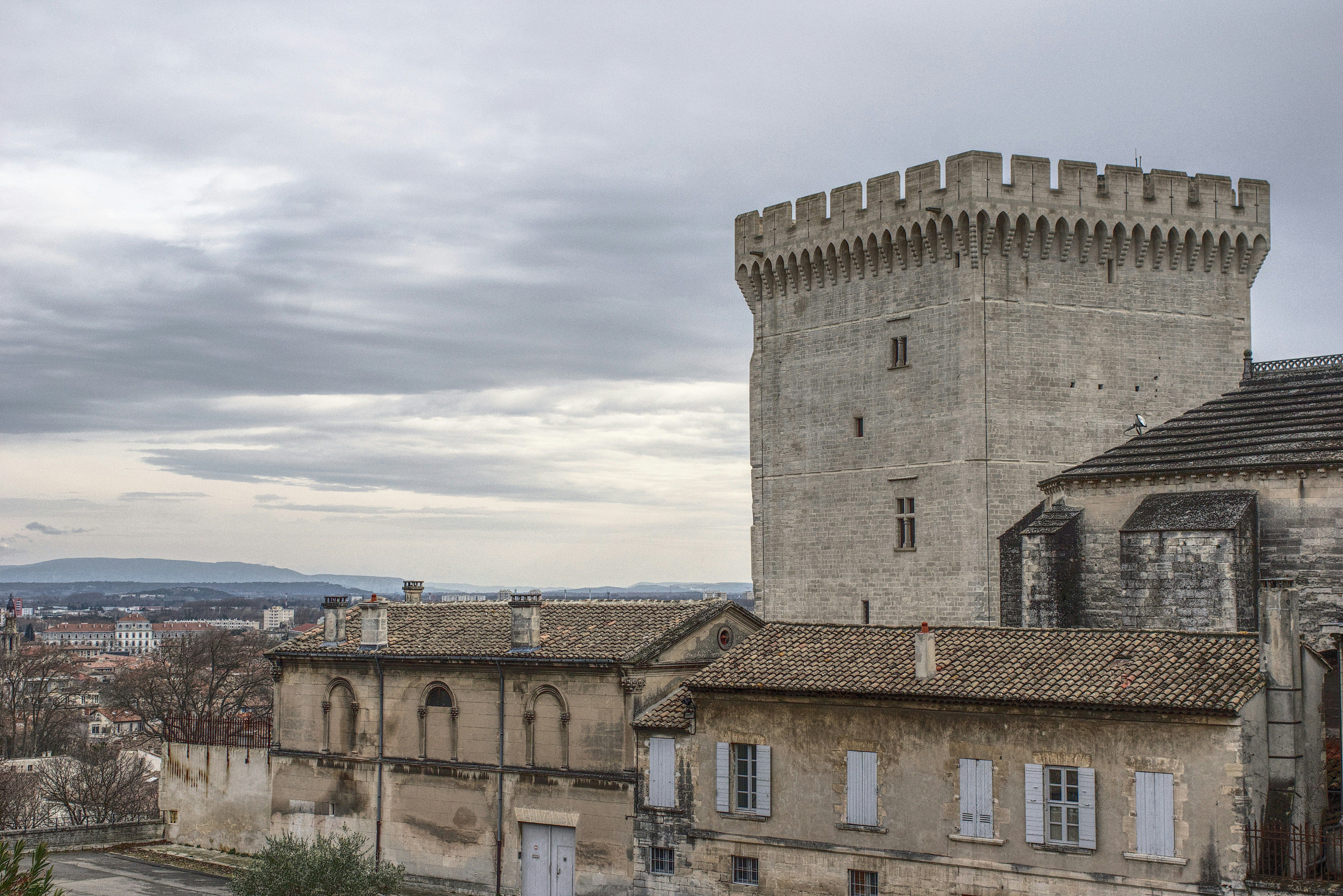 Der Papstpalast in Avignon. Man sieht einen mittelalterlichen Turm und Palast im Vordergrund, im Hintergrund den bewölkten Himmel.