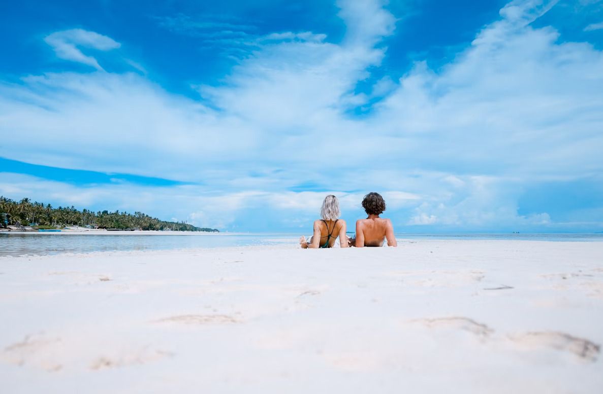 Dos personas tumbadas sobre la arena blanca mirando el mar bajo un cielo azul con nubes.