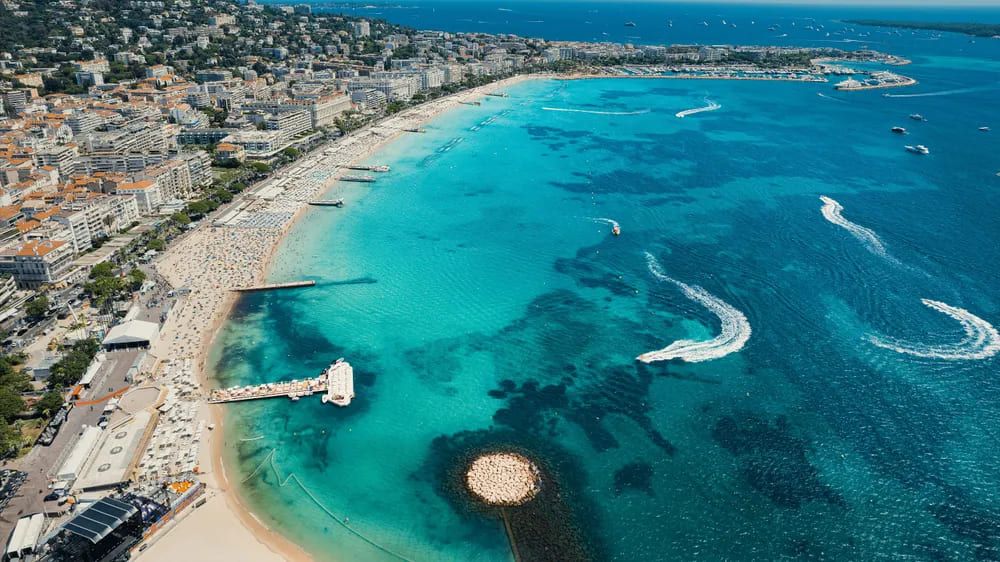 Vista aérea de la playa de La Croisette en Cannes, con aguas turquesas, bañistas y embarcaciones disfrutando del mar Mediterráneo.