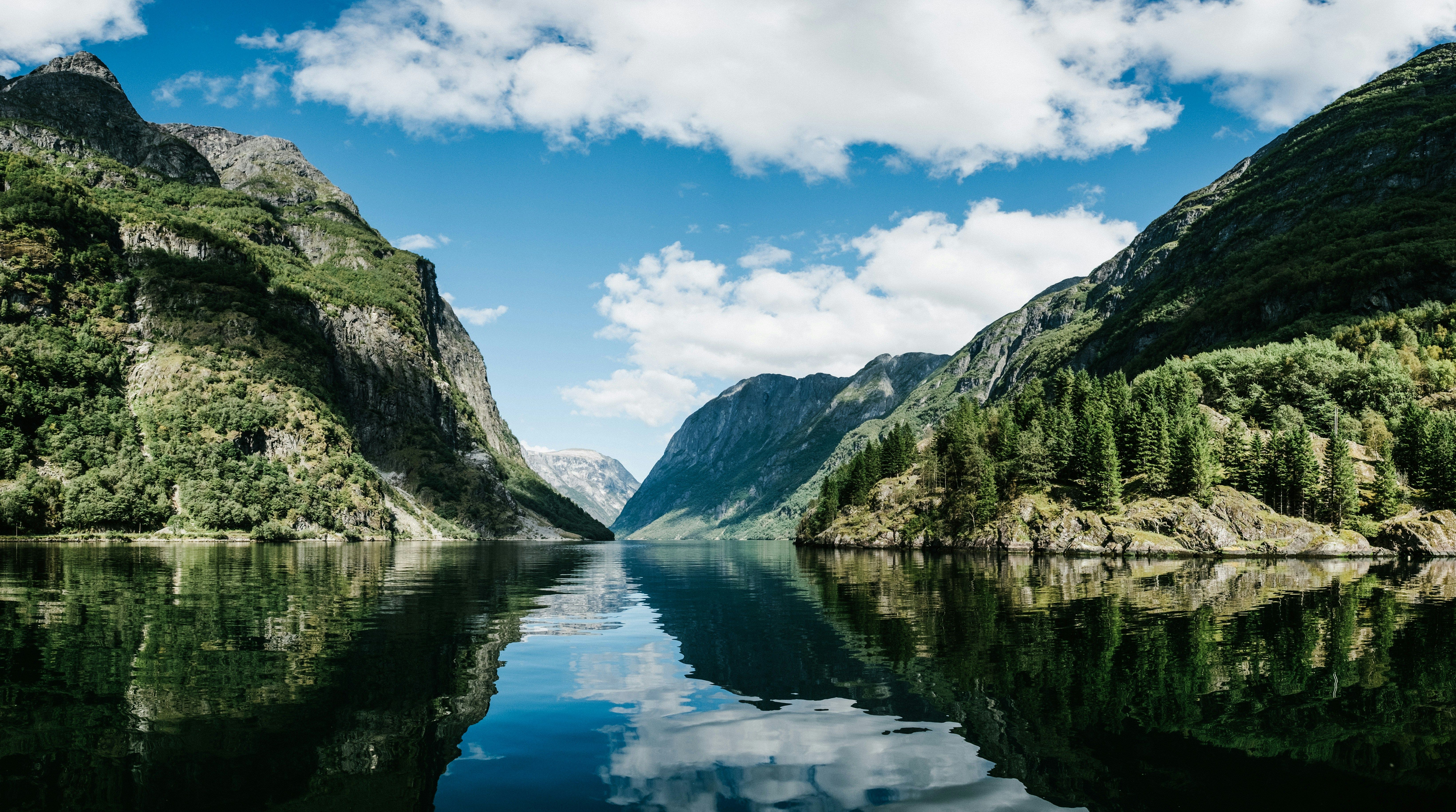 Ein beeindruckender Fjord in Norwegen. Links und rechts erheben sich gewaltige Bergketten, in der Mitte verläuft das blaue Meer, in dem sich der leicht bewölkte Himmel spiegelt.