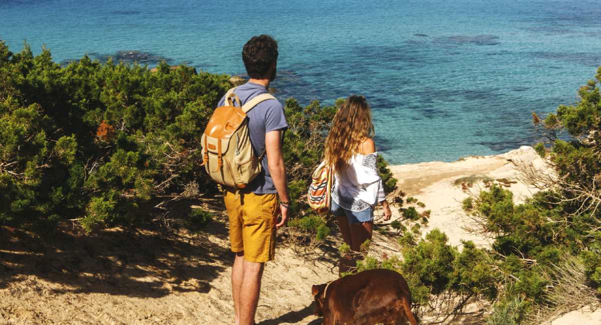 Pareja de excursionistas con mochilas y perro contemplando el mar en la costa de Mallorca