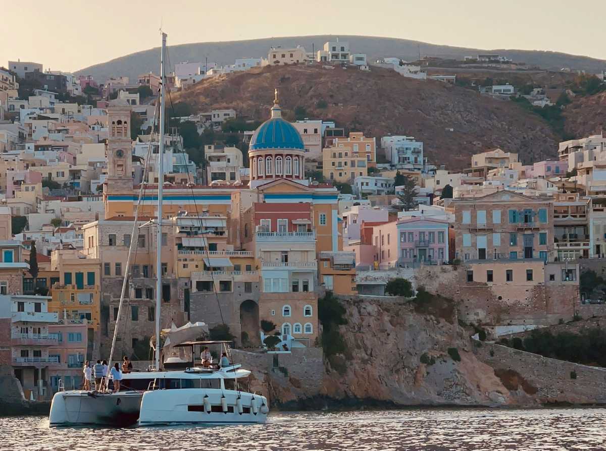Velero navegando frente a Ermoupoli, Syros, con casas de colores y la cúpula azul de la iglesia de San Nicolás al atardecer