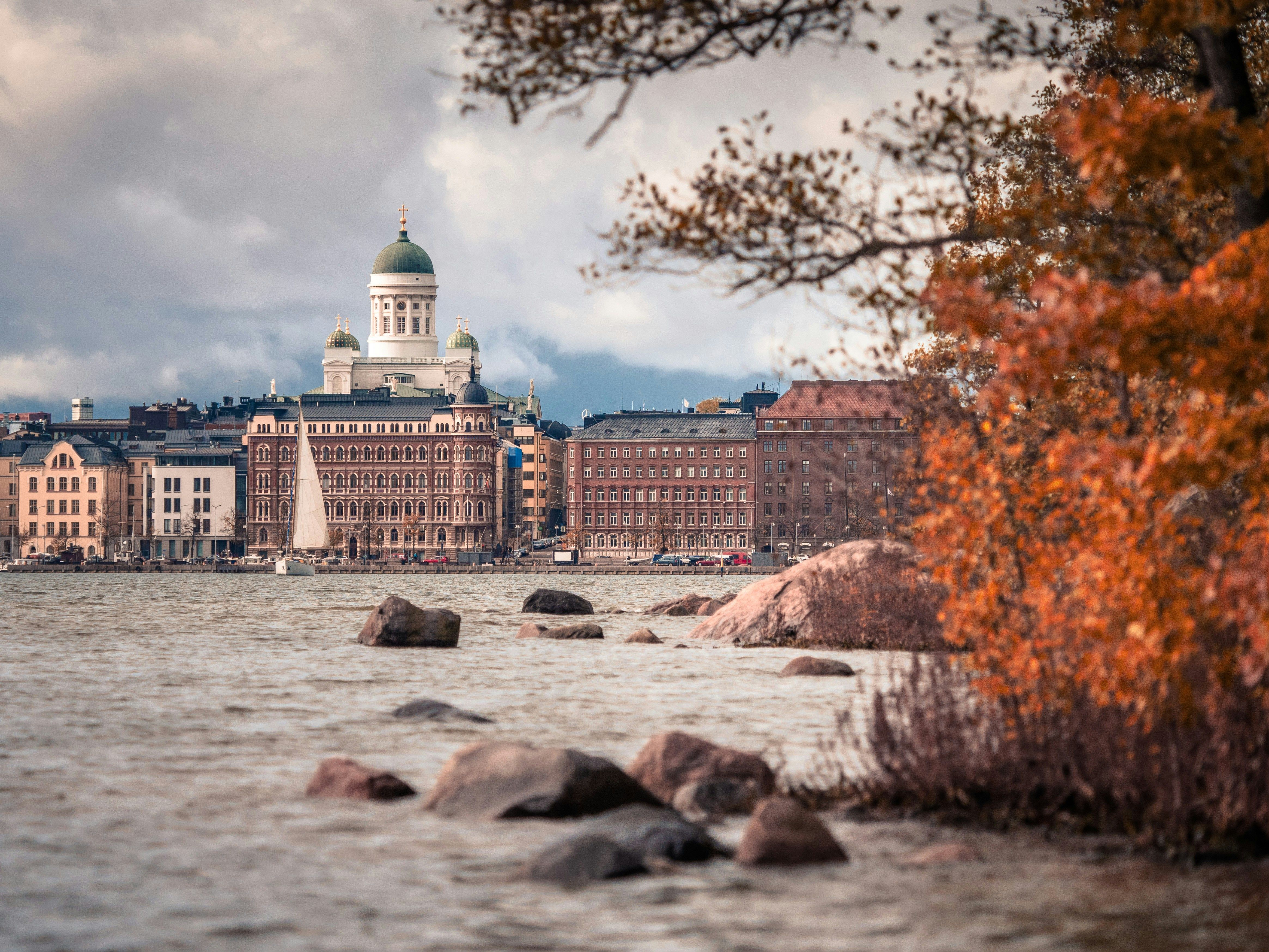 Blick auf die Skyline von Helsinki vom Wasser aus. Rechts im Bild sind Sträucher in herbstlichen Orange- und Rottönen. Der Himmel ist bewölkt.