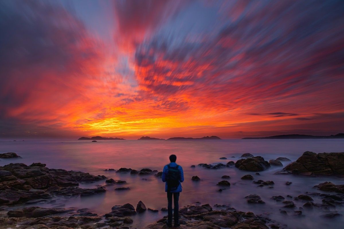 Persona contemplando el atardecer sobre las Islas Cíes desde la costa de Vigo, con cielo rojizo y mar en calma